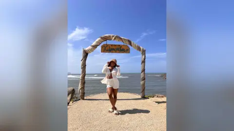 Stylish woman in white summer outfit and sun hat posing under a wooden arch at a sunny coastal beach destination.