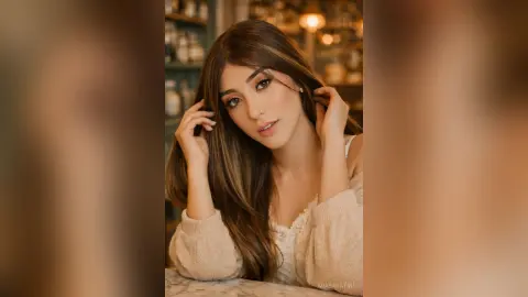 Young woman with long brown hair in a cozy white sweater posing in a warm, dimly lit cafe setting.