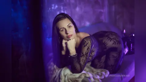 Woman in black lace bodysuit lounging on a sofa under moody purple lighting.