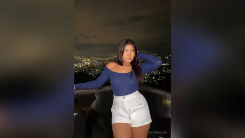 Young woman in a blue long-sleeve top and white shorts posing against a glowing city skyline at night.