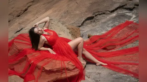 NataliaNacher lies on a rock wearing a long, flowing red dress with a high slit, showing her bare legs and feet.