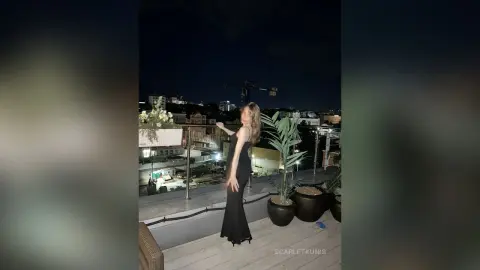 Elegant woman in a black evening gown standing on a rooftop terrace at night overlooking city lights.