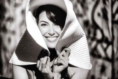 Smiling woman in a wide-brimmed sun hat posing against a patterned background in a bright, cinematic black and white style.