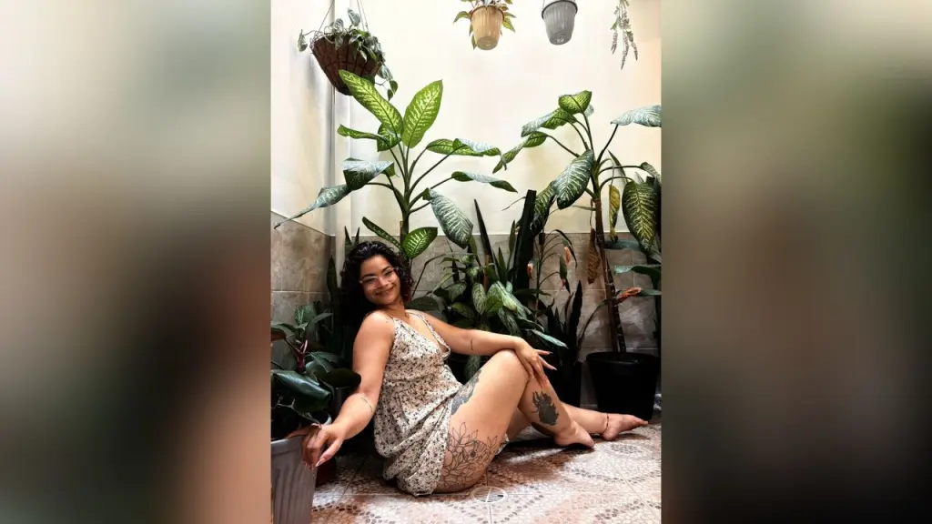 KasiaHill — Smiling woman in a floral sundress sitting on a tiled floor surrounded by lush green houseplants in a bright room.