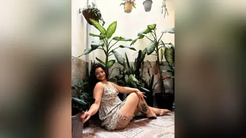 Smiling woman in a floral sundress sitting on a tiled floor surrounded by lush green houseplants in a bright room.