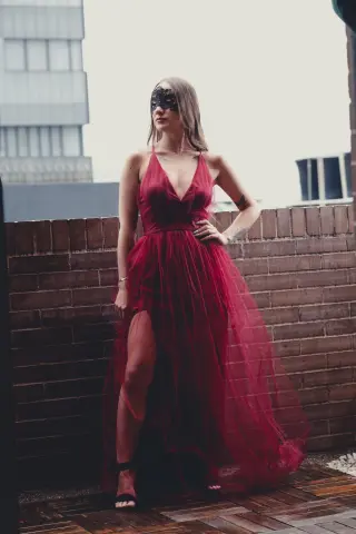 Elegant woman in a flowing red tulle gown and black masquerade mask posing on a rooftop at dusk.