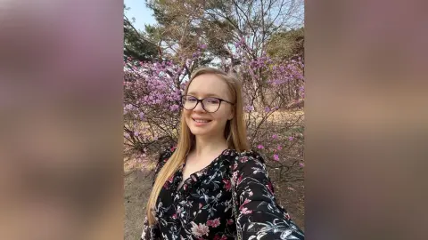 Smiling blonde woman in a floral blouse enjoying a sunny spring day among blooming pink flowers.
