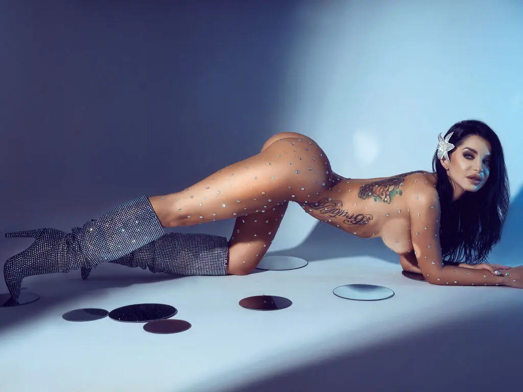 Glamorous woman with body jewels and tattoos lying on a studio floor in a moody, cinematic setting.