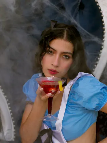 SusanaSanchez20 holds a red cocktail with a lemon twist, wearing a blue puff-sleeve top against a smoky, cobwebbed background.