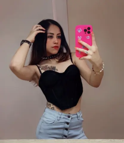 Young woman in a black corset top and light denim jeans taking a mirror selfie in a casual indoor setting.