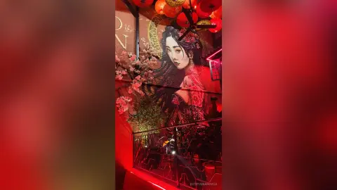 Elegant woman in a red dress standing before a large floral mural in a moody, red-lit restaurant setting.