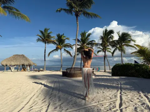Woman in a white crochet beach cover-up standing on a sunny tropical beach with palm trees and ocean view.