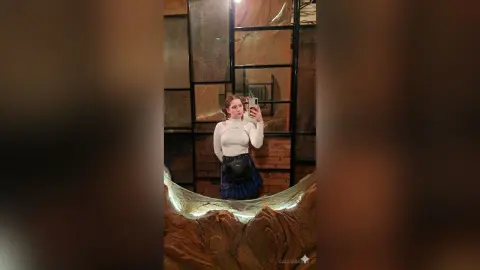 Young woman in a white turtleneck and blue skirt taking a mirror selfie in a modern, moody interior.