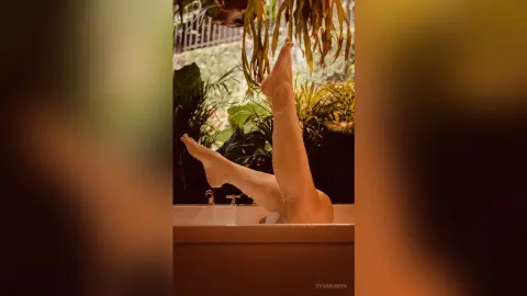 EvaMuskin lifts her bare legs high in the air while sitting in a bathtub surrounded by green plants.