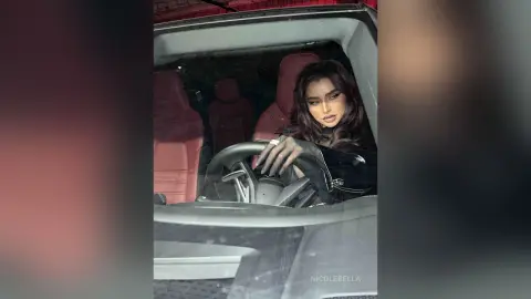 NicoleBella sits in the driver's seat of a car, looking toward the camera with long dark hair and heavy eye makeup.