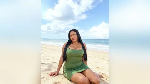 Young woman in a green summer dress sitting on a sunny beach with ocean waves in the background.