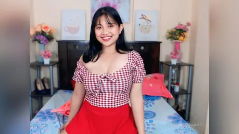 Smiling young woman in a red and white gingham top sitting on a bed in a cozy bedroom setting.