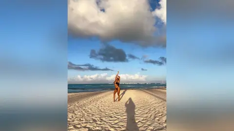 Alison stands naked on a sandy beach under a cloudy blue sky, posing with one arm raised above her head.