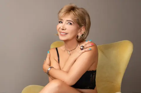 Smiling blonde woman in a black lace top sitting on a yellow velvet chair in a studio setting.