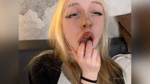 Blonde woman with freckles and nose ring in a cozy bedroom setting with a thoughtful expression.