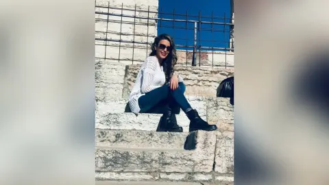 MontseFerrera sits on light stone steps wearing blue jeans, black boots, and a white lace top with sunglasses.