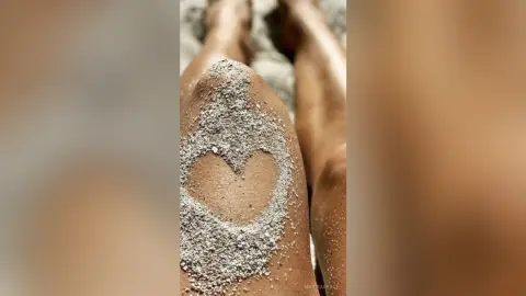 MarisaGrace shows a heart shape drawn in the sand on her thigh and leg at the beach.