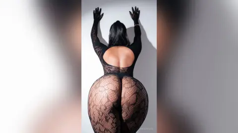 Curvy woman in a black sheer lace bodysuit posing against a white wall in a minimalist studio setting.