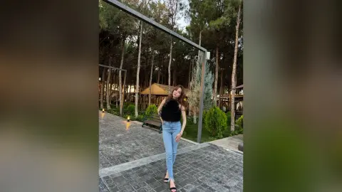 Stylish woman in light blue jeans and black fuzzy top posing in a serene outdoor forest garden setting.