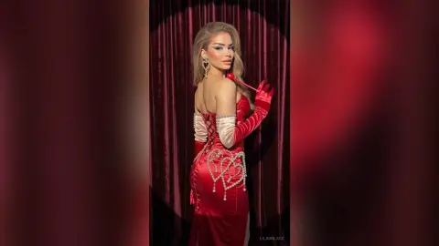 Glamorous woman in a red satin evening gown with pearl accents posing against red velvet curtains.