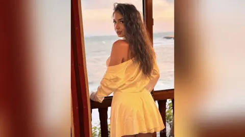 Smiling woman in a white off-the-shoulder dress relaxing on a balcony overlooking the ocean at sunset.