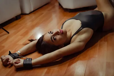 Young woman in a black bodysuit lying on a wooden floor wearing leather wrist cuffs in a moody setting.