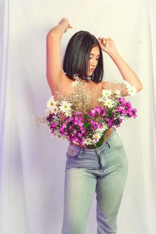 Young woman in denim jeans holding a large bouquet of pink and white wildflowers against a white backdrop.