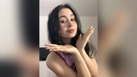 MinnieMile poses with her hands near her face, wearing a pink and red top with her long dark hair down.