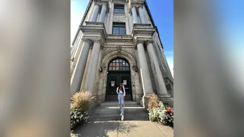 Stylish woman in white top and jeans standing before a grand classical stone building under a clear blue sky.