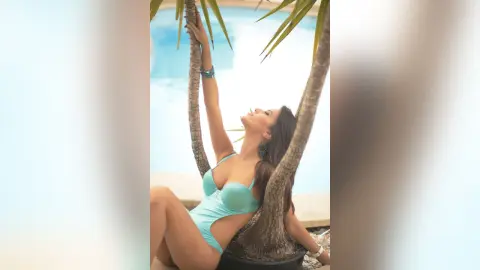 MontseFerrera poses in a light blue bodysuit while leaning back against a palm tree by a swimming pool.