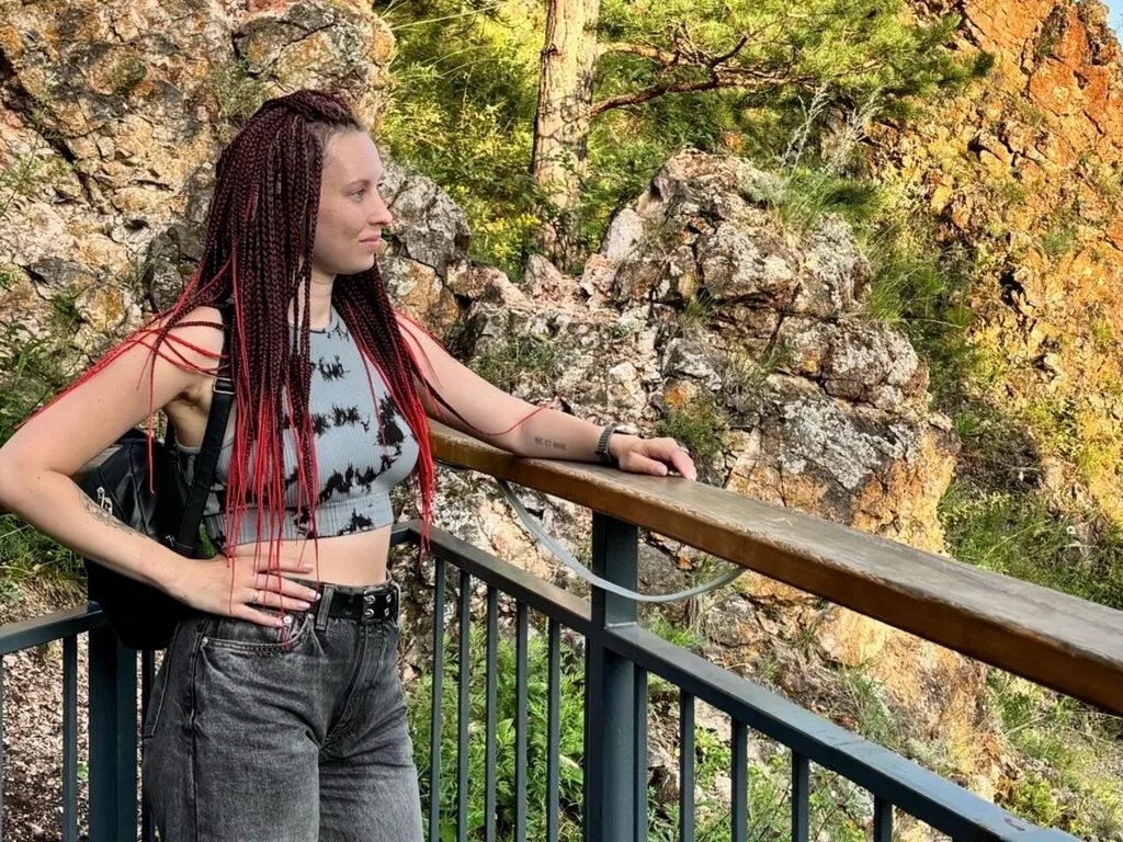 AmeliaMeds with long red braids stands on a balcony overlooking a rocky mountain landscape.