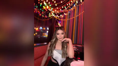 Young woman in a white strapless top smiling under colorful festive string lights in a vibrant lounge setting.