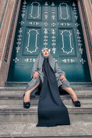 Elegant woman in a black abaya and grey hijab sitting on stone steps in front of ornate teal doors.