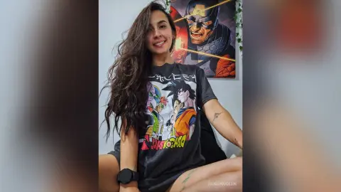 RossiNoleen smiles while wearing a dark grey anime graphic t-shirt and sitting against a white wall near a framed poster.