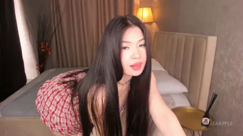 Young woman with long dark hair in a red plaid shirt relaxing in a cozy bedroom setting.