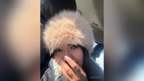 Smiling young woman in a cozy beige faux fur hat and winter jacket sitting in a car on a sunny day.