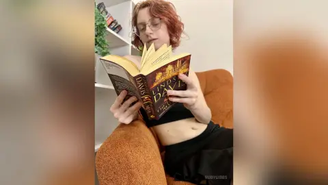 Red-haired woman in a black crop top reading a fantasy novel while relaxing in a cozy orange armchair.