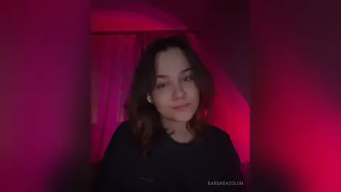 Young woman with wavy hair in a dark room illuminated by moody pink ambient lighting.