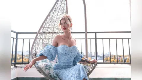 DaisyLin sits in a hanging wicker chair wearing a light blue off-the-shoulder lace dress with blonde hair and red lipstick.