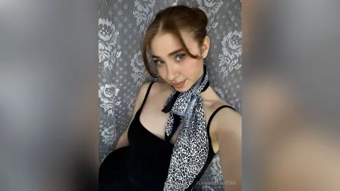 Young woman in a black dress and leopard print neck scarf posing against a floral curtain backdrop.