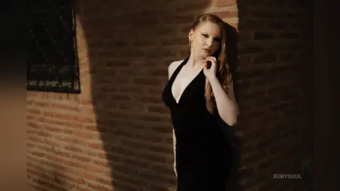 RubySoul poses against a brick wall wearing a low-cut black dress that shows off her cleavage and shoulders.