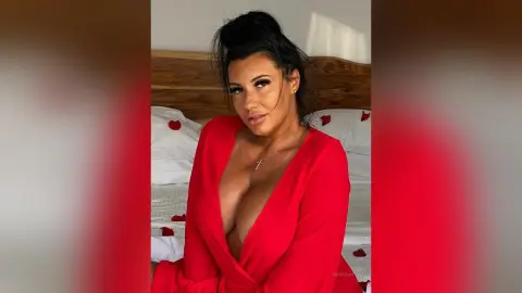 MissyJolie poses in a red wrap top with a deep cleavage, looking at the camera while sitting on a bed with white sheets.