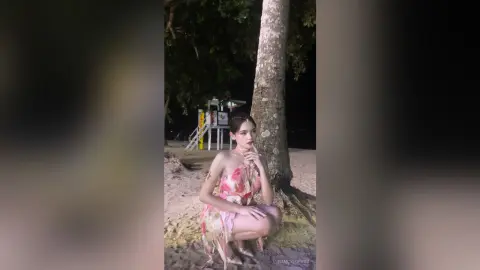 NancyLoppez squats on a sandy beach at night wearing a pink floral halter-neck dress.