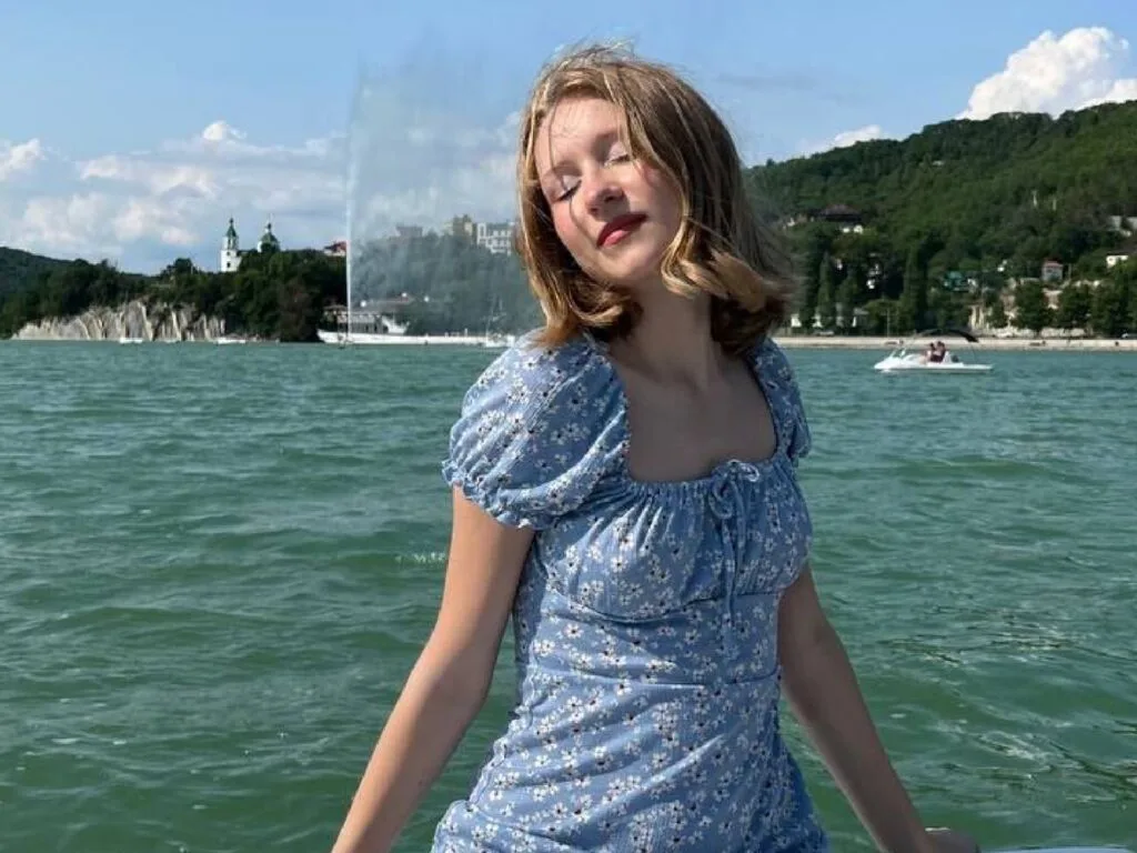MozelleEzzo wears a blue floral sundress while posing outdoors by the water on a sunny day.
