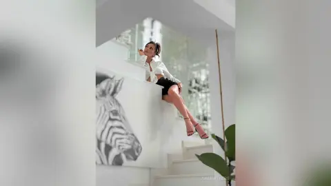 AnaMathers sits on white stairs wearing a white button-down shirt and black shorts.